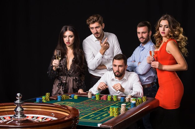 Group of young people behind roulette table on black background. Young people made bets in the game and wait for the result. Bright emotions