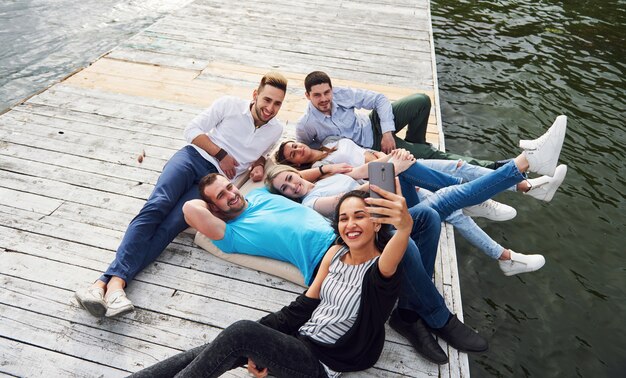 A group of young people sitting on the edge of the pier, and make selfie. Friends enjoying a game on the lake.