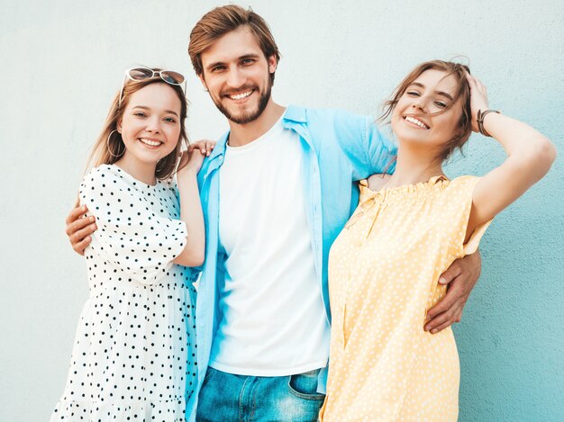 通りでポーズをとる若い3人のスタイリッシュな友人のグループ。ファッション男とカジュアルな夏服を着た2人のかわいい女の子。壁の近くで楽しんでいる笑顔のモデル。陽気な女性と屋外の男