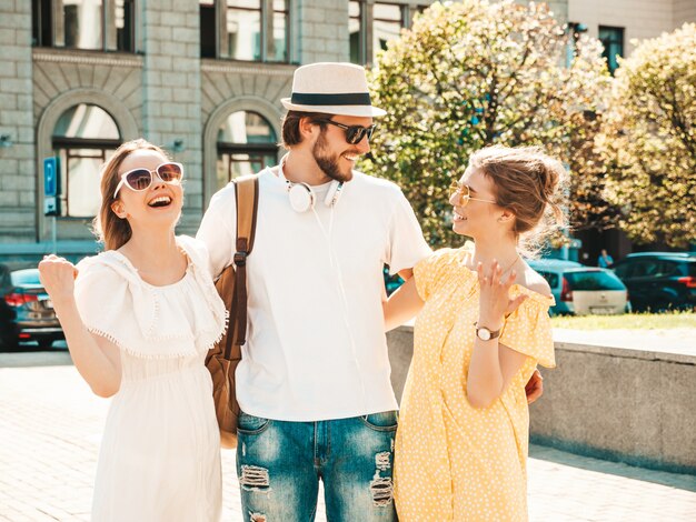 通りでポーズをとる若い3人のスタイリッシュな友人のグループ。ファッション男とカジュアルな夏服を着た2人のかわいい女の子。サングラスで楽しい笑顔のモデル。陽気な女性とチャットの男