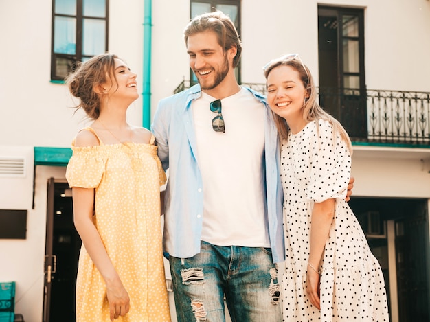 Group of young three stylish friends posing in the street. Fashion man and two cute girls dressed in casual summer clothes. Smiling models having fun in sunglasses.Cheerful women and guy chatting