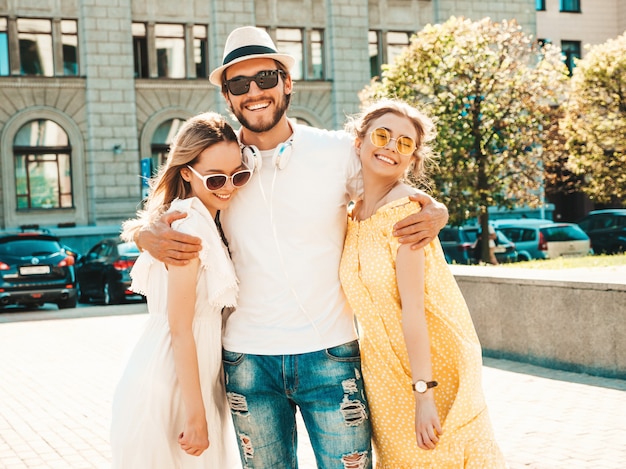 通りでポーズをとる若い3人のスタイリッシュな友人のグループ。ファッション男とカジュアルな夏服を着た2人のかわいい女の子。サングラスで楽しんでいる笑顔のモデル。陽気な女性と男が夢中になる