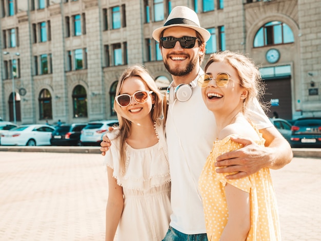 Group of young three stylish friends posing in the street. Fashion man and two cute girls dressed in casual summer clothes. Smiling models having fun in sunglasses.Cheerful women and guy going crazy