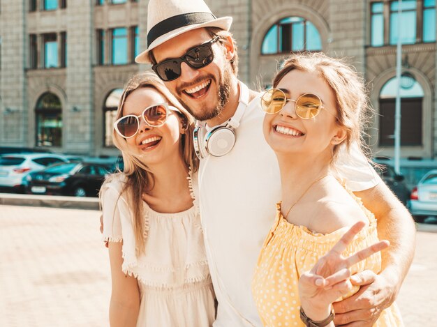 通りでポーズをとる若い3人のスタイリッシュな友人のグループ。ファッション男とカジュアルな夏服を着た2人のかわいい女の子。サングラスで楽しんでいる笑顔のモデル。陽気な女性と男が夢中になる