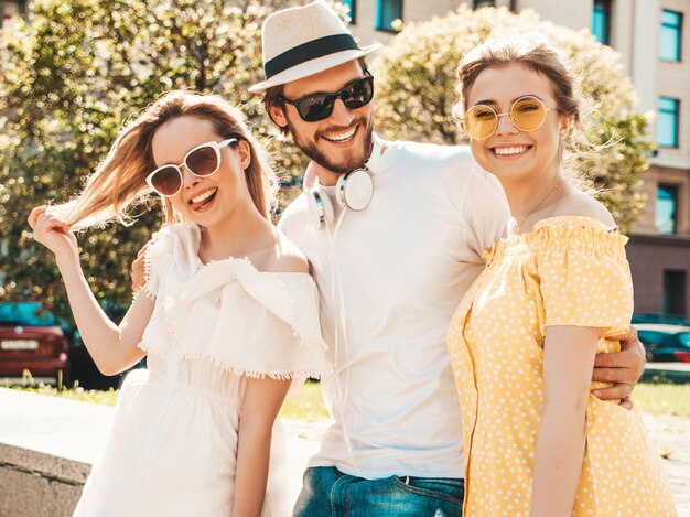 通りでポーズをとる若い3人のスタイリッシュな友人のグループ。ファッション男とカジュアルな夏服を着た2人のかわいい女の子。サングラスで楽しんでいる笑顔のモデル。陽気な女性と男が夢中になる