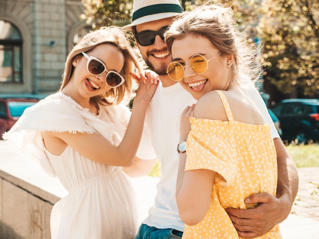 通りでポーズをとる若い3人のスタイリッシュな友人のグループ。ファッション男とカジュアルな夏服を着た2人のかわいい女の子。サングラスで楽しんでいる笑顔のモデル。陽気な女性と男が夢中になる