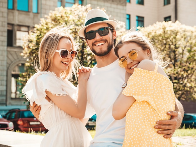 通りでポーズをとる若い3人のスタイリッシュな友人のグループ。ファッション男とカジュアルな夏服を着た2人のかわいい女の子。サングラスで楽しんでいる笑顔のモデル。陽気な女性と男が夢中になる