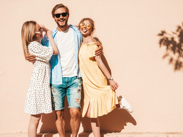 通りでポーズをとる若い3人のスタイリッシュな友人のグループ。ファッション男とカジュアルな夏服を着た2人のかわいい女の子。サングラスで楽しんでいる笑顔のモデル。陽気な女性と男が夢中になる