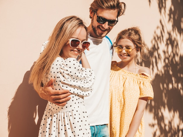 通りでポーズをとる若い3人のスタイリッシュな友人のグループ。ファッション男とカジュアルな夏服を着た2人のかわいい女の子。サングラスで楽しんでいる笑顔のモデル。陽気な女性と男が夢中になる