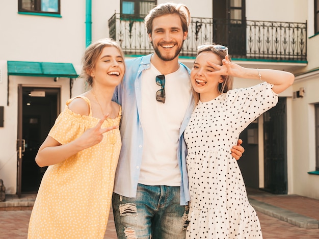 Group of young three stylish friends posing in the street. Fashion man and two cute girls dressed in casual summer clothes. Smiling models having fun in sunglasses.Cheerful women and guy going crazy