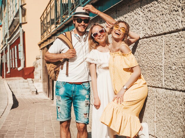 通りでポーズをとる若い3人のスタイリッシュな友人のグループ。ファッション男とカジュアルな夏服を着た2人のかわいい女の子。サングラスで楽しい笑顔のモデル。陽気な女性と屋外の男