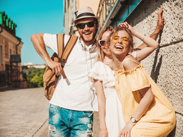 通りでポーズをとる若い3人のスタイリッシュな友人のグループ。ファッション男とカジュアルな夏服を着た2人のかわいい女の子。サングラスで楽しい笑顔のモデル。陽気な女性と屋外の男