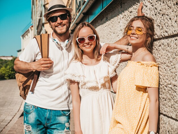 Group of young three stylish friends posing in the street. Fashion man and two cute girls dressed in casual summer clothes. Smiling models having fun in sunglasses.Cheerful women and guy outdoors