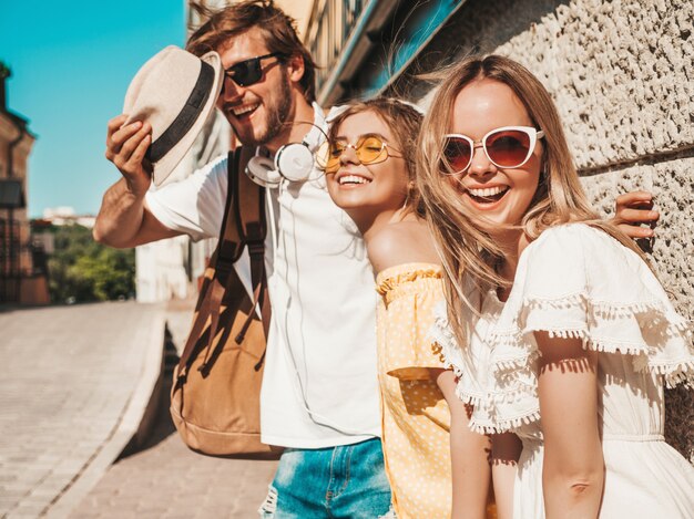 通りでポーズをとる若い3人のスタイリッシュな友人のグループ。ファッション男とカジュアルな夏服を着た2人のかわいい女の子。サングラスで楽しい笑顔のモデル。陽気な女性と屋外の男