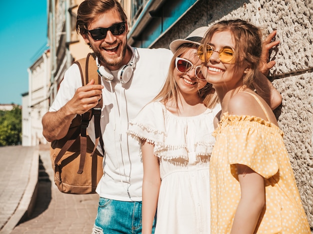 Group of young three stylish friends posing in the street. Fashion man and two cute girls dressed in casual summer clothes. Smiling models having fun in sunglasses.Cheerful women and guy outdoors