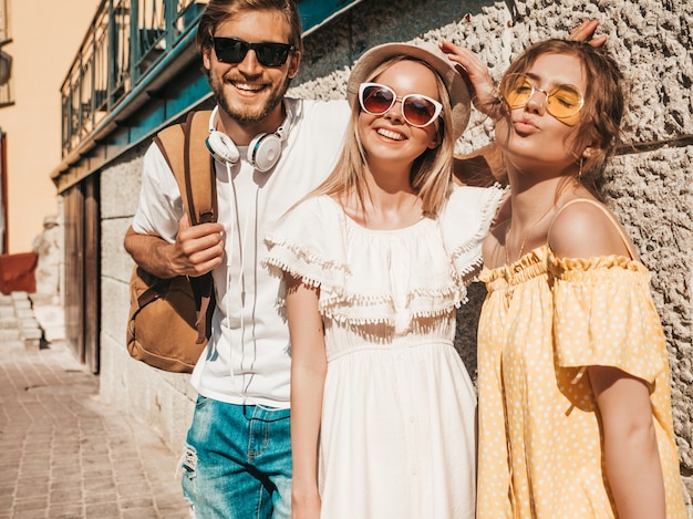 Group of young three stylish friends posing in the street. Fashion man and two cute girls dressed in casual summer clothes. Smiling models having fun in sunglasses.Cheerful women and guy outdoors