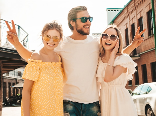 通りでポーズをとる若い3人のスタイリッシュな友人のグループ。ファッション男とカジュアルな夏服を着た2人のかわいい女の子。サングラスで楽しい笑顔のモデル。陽気な女性と屋外の男