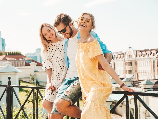 Group of young three stylish friends posing in the street. Fashion man and two cute girls dressed in casual summer clothes. Smiling models having fun in sunglasses.Cheerful women and guy outdoors