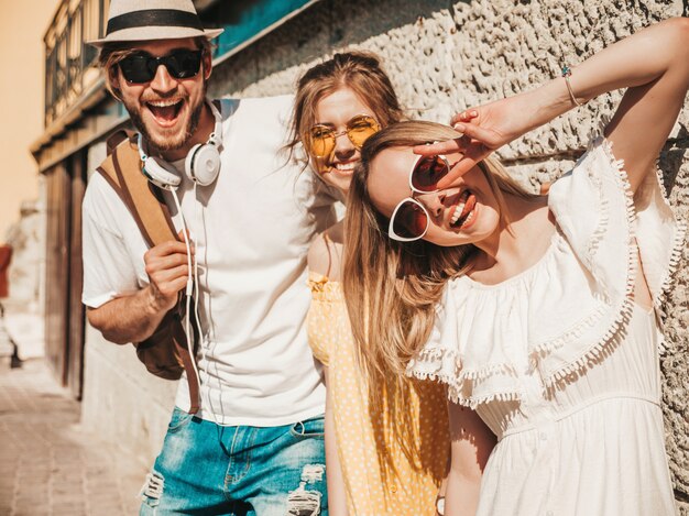 通りでポーズをとる若い3人のスタイリッシュな友人のグループ。ファッション男とカジュアルな夏服を着た2人のかわいい女の子。サングラスで楽しい笑顔のモデル。陽気な女性と男は舌を示す