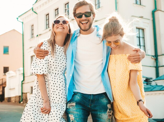 通りでポーズをとる若い3人のスタイリッシュな友人のグループ。ファッション男とカジュアルな夏服を着た2人のかわいい女の子。サングラスで楽しい笑顔のモデル。陽気な女性とサスネットで男