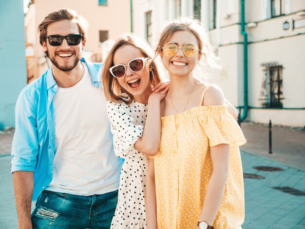 Group of young three stylish friends posing in the street. Fashion man and two cute girls dressed in casual summer clothes. Smiling models having fun in sunglasses.Cheerful women and guy at susnet