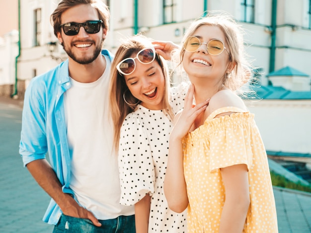 Group of young three stylish friends posing in the street. Fashion man and two cute girls dressed in casual summer clothes. Smiling models having fun in sunglasses.Cheerful women and guy at susnet