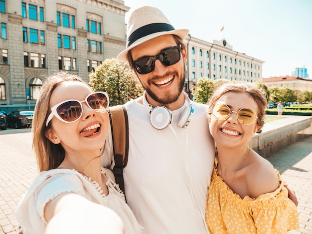 Group of young three stylish friends in the street.Man and two cute girls dressed in casual summer clothes.Smiling models having fun in sunglasses.Women and guy making photo selfie on smartphone