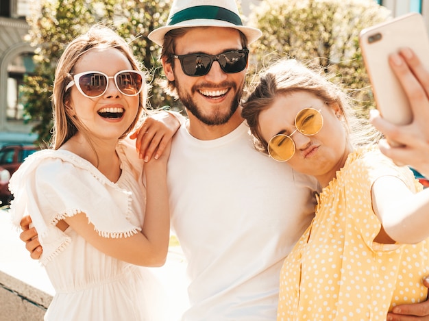 Group of young three stylish friends in the street.Man and two cute girls dressed in casual summer clothes.Smiling models having fun in sunglasses.Women and guy making photo selfie on smartphone
