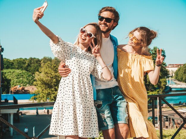 通りでスタイリッシュな若い3人の友人のグループ。男と2人のかわいい女の子がカジュアルな夏服に身を包んだ。サングラスで楽しい笑顔のモデル。スマートフォンで写真を撮る女性と男