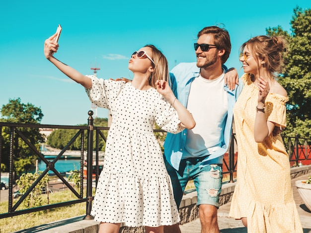 Group of young three stylish friends in the street.man and two cute girls dressed in casual summer clothes.smiling models having fun in sunglasses.women and guy making photo selfie on smartphone