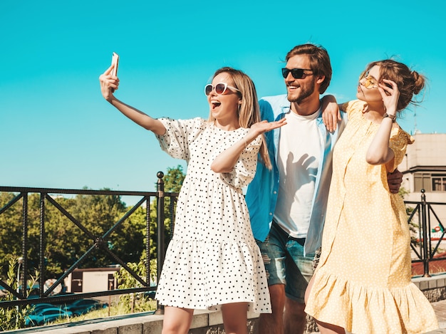 通りでスタイリッシュな若い3人の友人のグループ。男と2人のかわいい女の子がカジュアルな夏服に身を包んだ。サングラスで楽しい笑顔のモデル。スマートフォンで写真を撮る女性と男