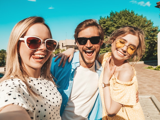 Group of young three stylish friends in the street.Man and two cute girls dressed in casual summer clothes.Smiling models having fun in sunglasses.Women and guy making photo selfie on smartphone