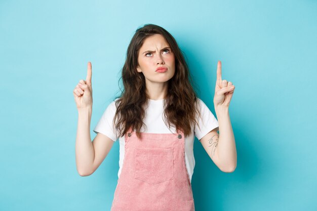 Grumpy cute brunette girl frowning displeased, sulking and looking disappointed up, pointing fingers on top, standing against blue background