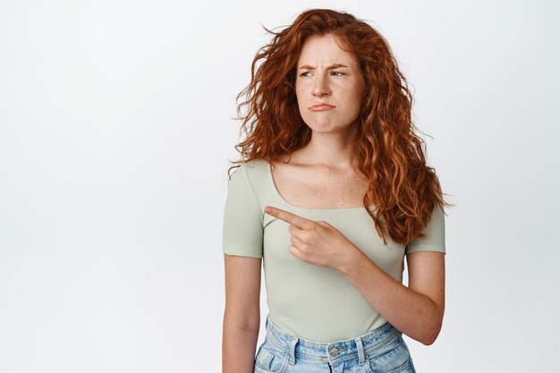 Grumpy redhead woman pointing and looking left with pouting face expression wearing tshirt and jeans white background