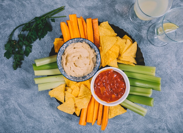 Guacamole and salsa sauce in bowl with carrot; celery stem; juice; coriander and tortilla chips over concrete background