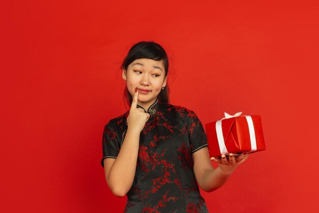 선물이 뭔지 맞춰보세요. 해피 중국 설날. 빨간색 배경에 고립 된 아시아 젊은 여자의 초상화. 전통 옷을 입은 여성 모델이 행복해 보입니다. 축하, 휴일, 감정. Copyspace.