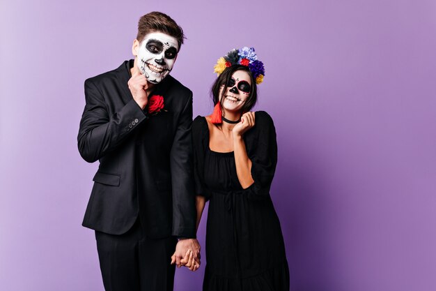 Guilty boyfriend and his girlfriend are trying to smile sweetly. Portrait of woman with bright accessories and man in dark classic suit posing with makeup for Halloween.