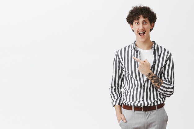 Guy being excited before making new tattoo yelling from delight and happiness standing amused and thrulled in striped cool shirt pointing at upper left corner posing over gray wall