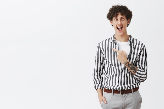 Guy being excited before making new tattoo yelling from delight and happiness standing amused and thrulled in striped cool shirt pointing at upper left corner posing over gray wall
