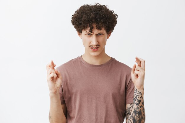 Guy believes in power of mind. Intense serious-looking determined and ambitious guy with curly hairstyle and tattoos hoping get sholarship crossing fingers for good luck and staring strict