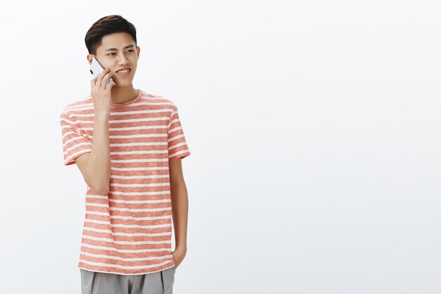 Guy calling friend talking casually via smartphone standing to left side of copy space gazing aside with nice smile using cellphone to connect with family while living abroad over white wall