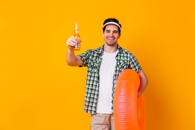 隔離されたスペースにビールとオレンジ色の膨脹可能な円のボトルを保持しているキャップと白いTシャツの男。