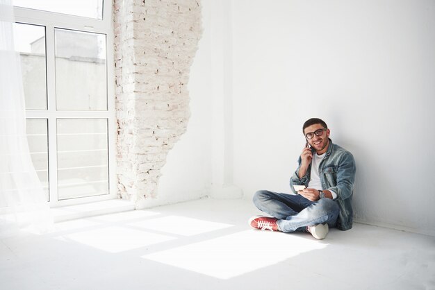 A guy in casual clothes is sitting at home in an empty apartment holding a credit card and calling on the phone.