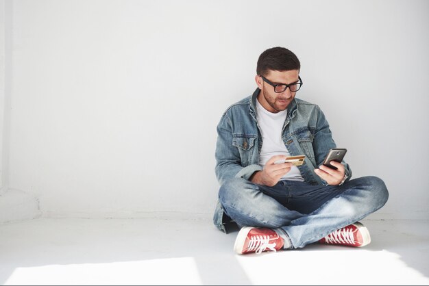 A guy in casual clothes is sitting at home in an empty apartment holding a credit card