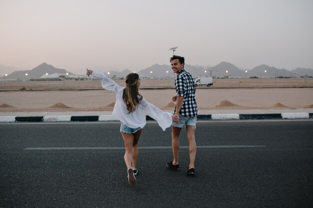 Guy in denim shorts and long-haired woman in trendy blouse running across the road and enjoys mountain view. Laughing young couple holding hands walking on highway and having fun outside in summer