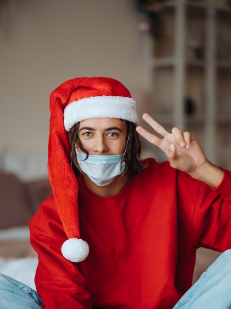 Парень в маске и шляпе Санта-Клауса позирует и смотрит в камеру дома