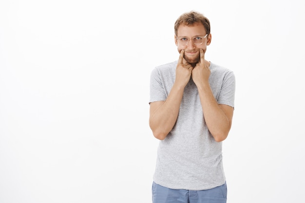 Guy fed up pretending he okay pulling happy smile on face but feeling gloomy and unhappy stretching grin with fingers on lips corners staring indifferent and careless