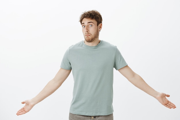 Guy feeling confused after loosing wallet. Portrait of questioned timid attractive man in t-shirt, spreading palms and shrugging with awkward unaware expression