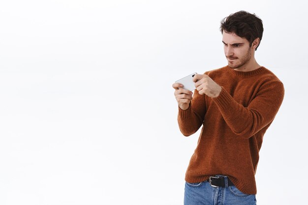 Парень сосредоточился на жестком уровне Серьезно выглядящий молодой красивый мужчина в повседневной одежде держит смартфон горизонтально, хмурится и смотрит на мобильный дисплей, играя в интересную игру на белом фоне