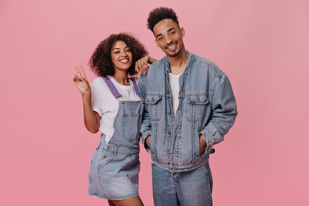 Guy and girl in denim outfits showing peace sign on pink wall